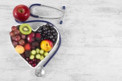 Heart health, and cholesterol diet concept. Healthy fruits in heart shaped bowl with stethoscope and green apple on white vintage wooden table.