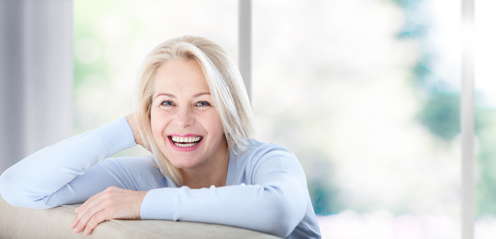 Active beautiful middle-aged woman smiling friendly and looking in camera near window at home. Woman's face closeup. Selective focus.