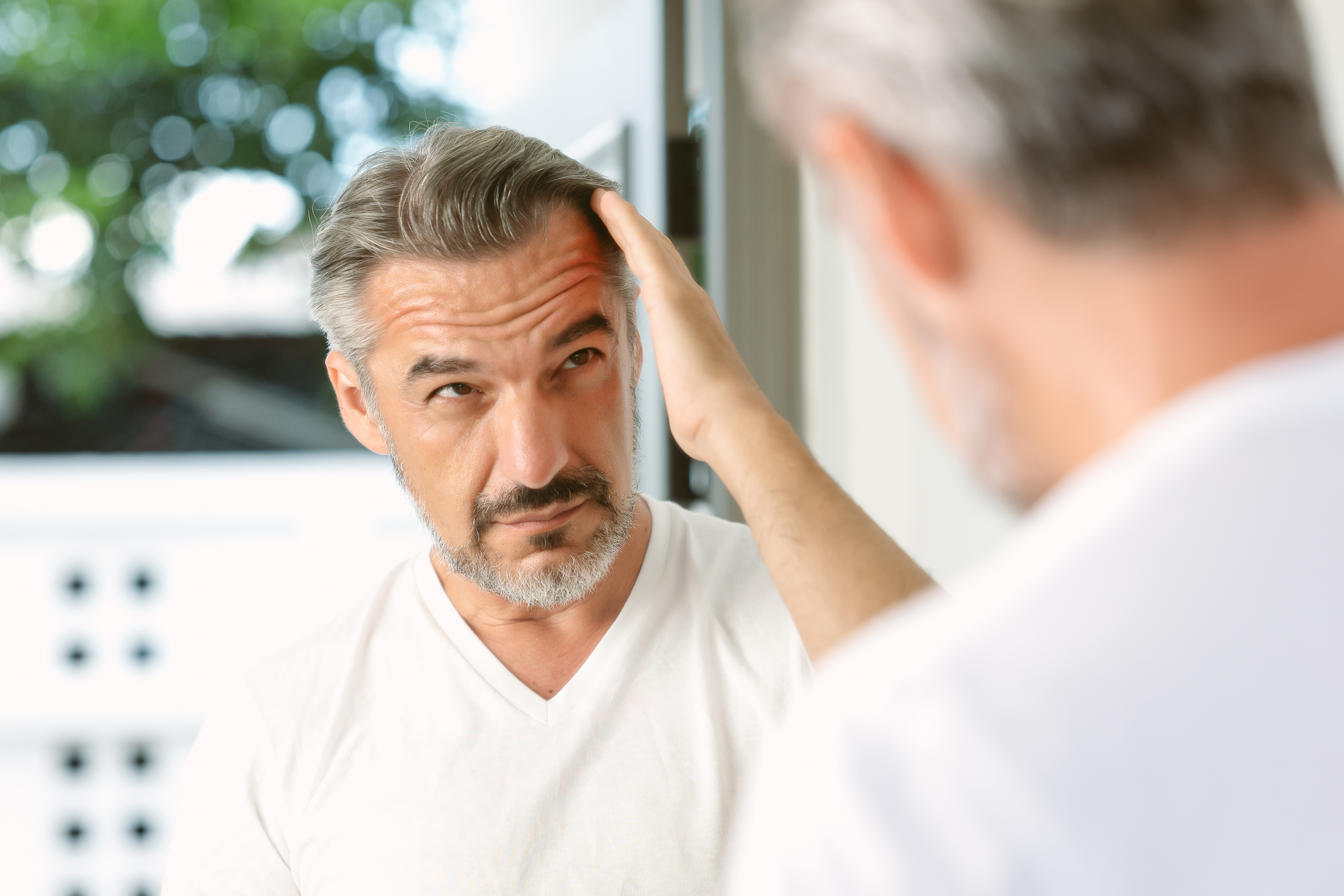 Handsome middle age caucasian man with beard looks in the mirror. Middle-aged man concerned by hair loss. Men’s personal care. Morning routine. Receding hairline, haircare, beauty concept