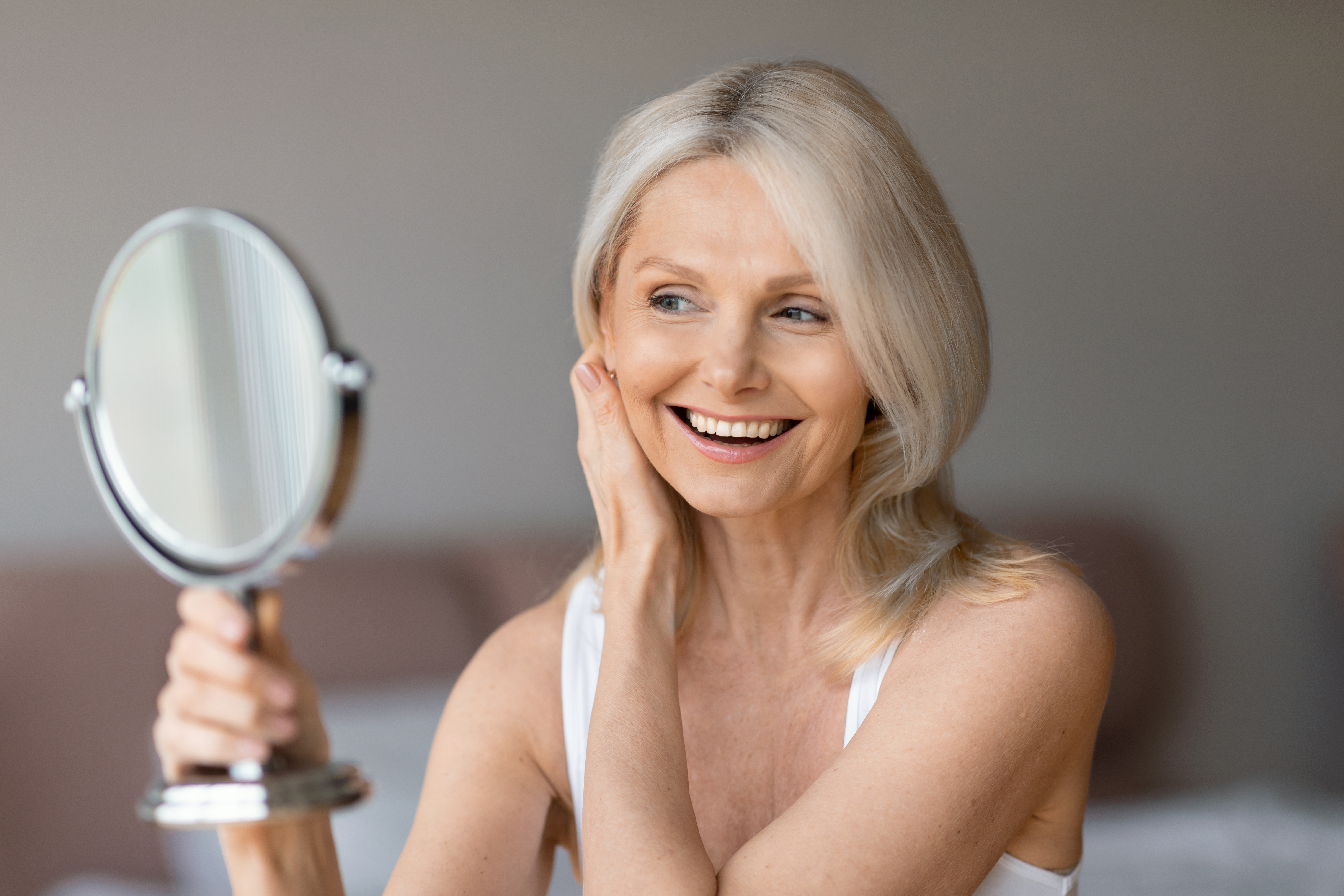 Aged natural beauty concept. Happy senior woman looking at mirror, touching her face and smiling to reflection
