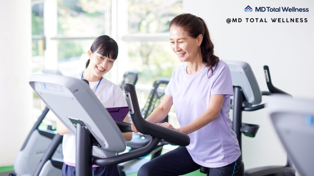 A smiling woman in purple workout clothes exercises on gym equipment in Brea wellness center with a fitness trainer